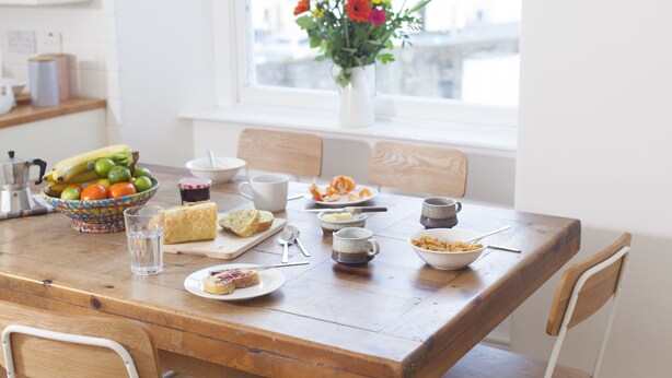 Una mesa de comedor con diversos alimentos y bebidas, un florero con flores, y una ventana con vista a edificios.