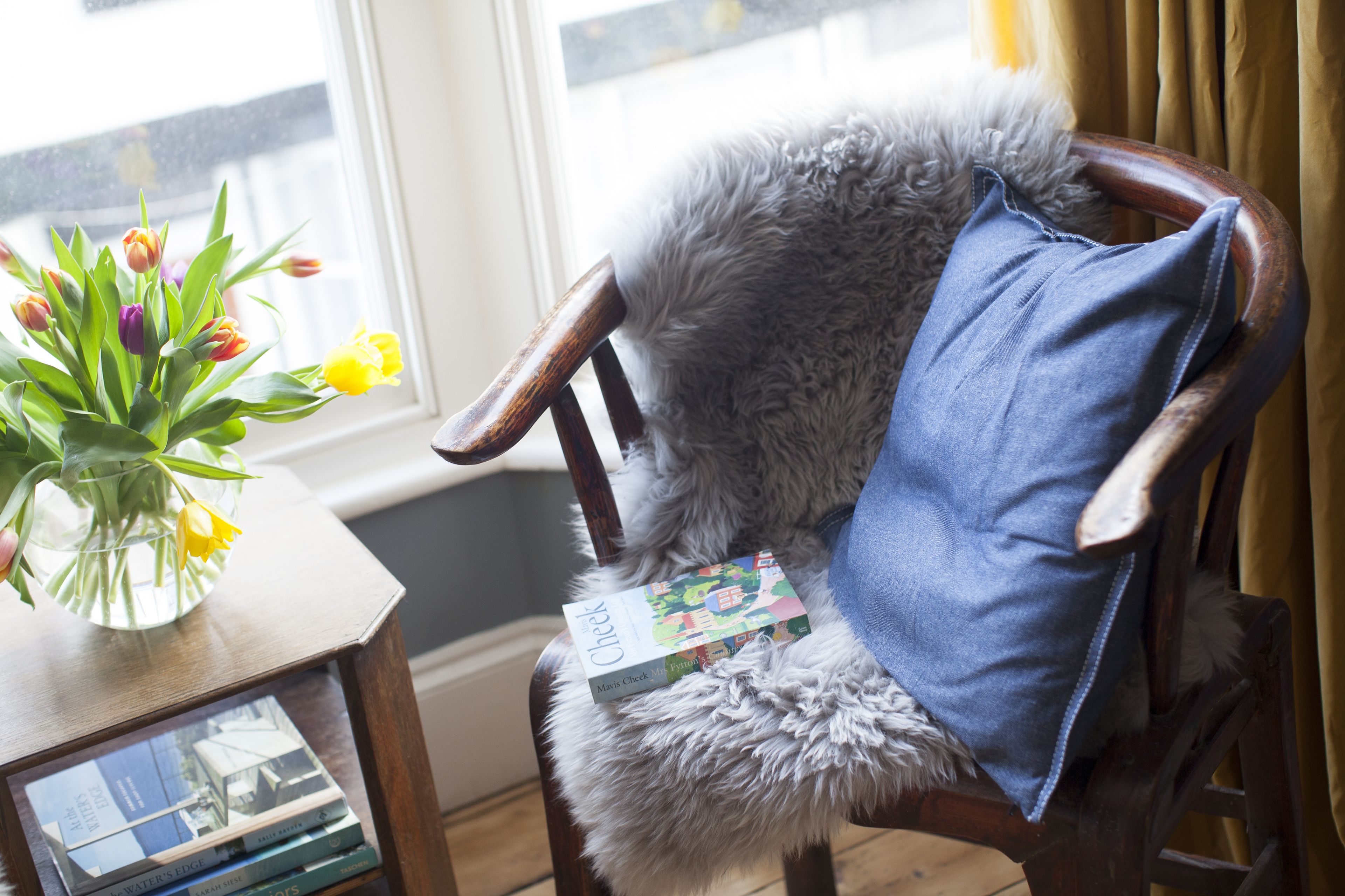 En stol med en mjuk, grå sittkudde och en blå prydnadskudde, en vas med gula blommor och en bok med titeln ”Check” på ett litet träbord.