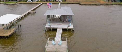 A waterfront property with a dock extending into a murky body of water. There are several buildings with distinct architectural styles, including a house with a red roof and another with a white exterior. A flag is visible on the dock, and the area is surrounded by vegetation and a fence.