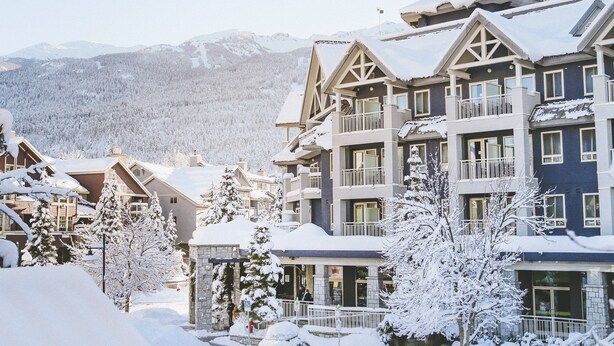 A multi-story hotel with a snow-covered roof and balconies, surrounded by snow-laden trees and mountains in the background.