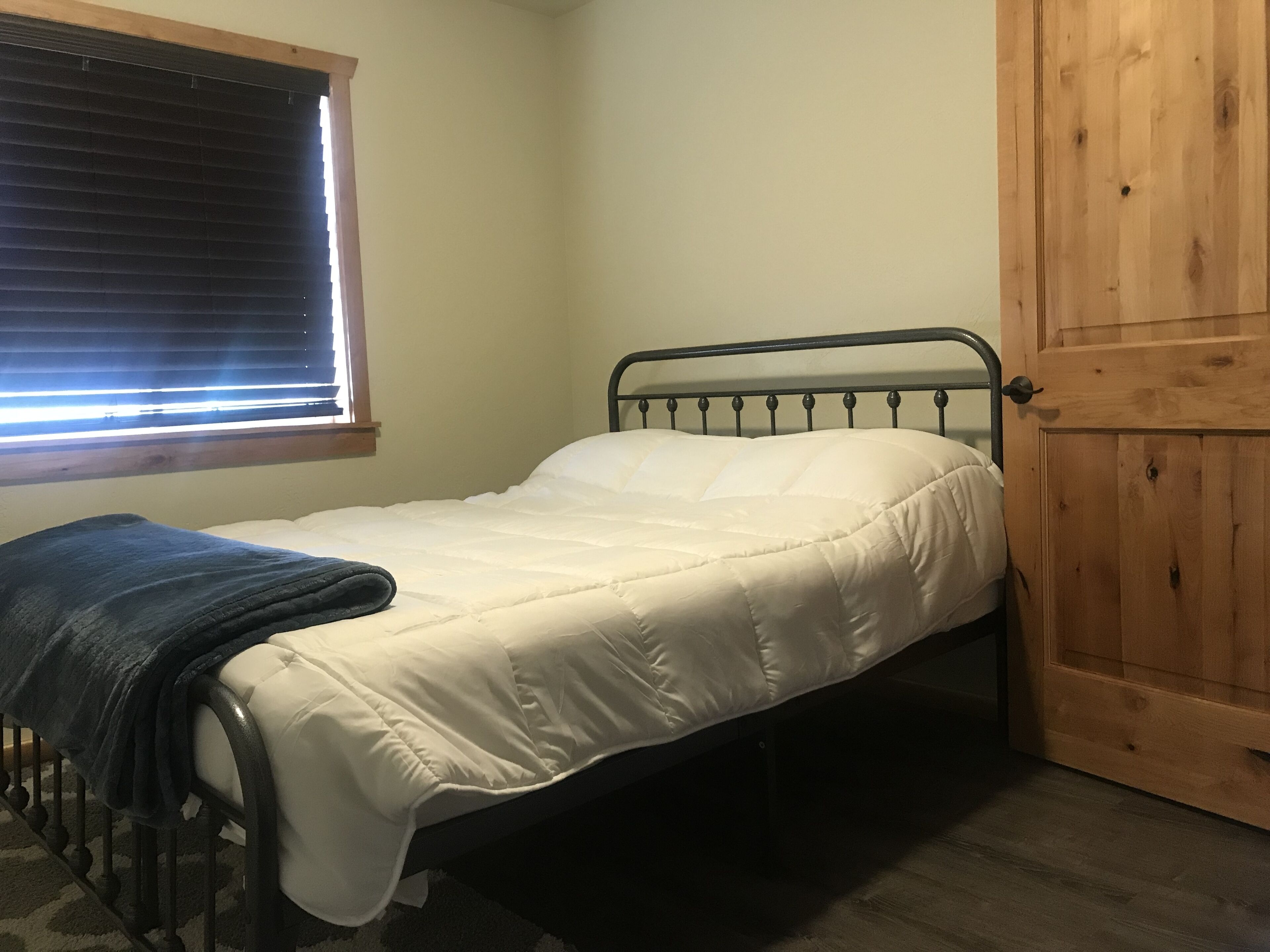 Un lit simple avec un matelas blanc et une couverture foncée, une porte en bois et une fenêtre avec des stores.