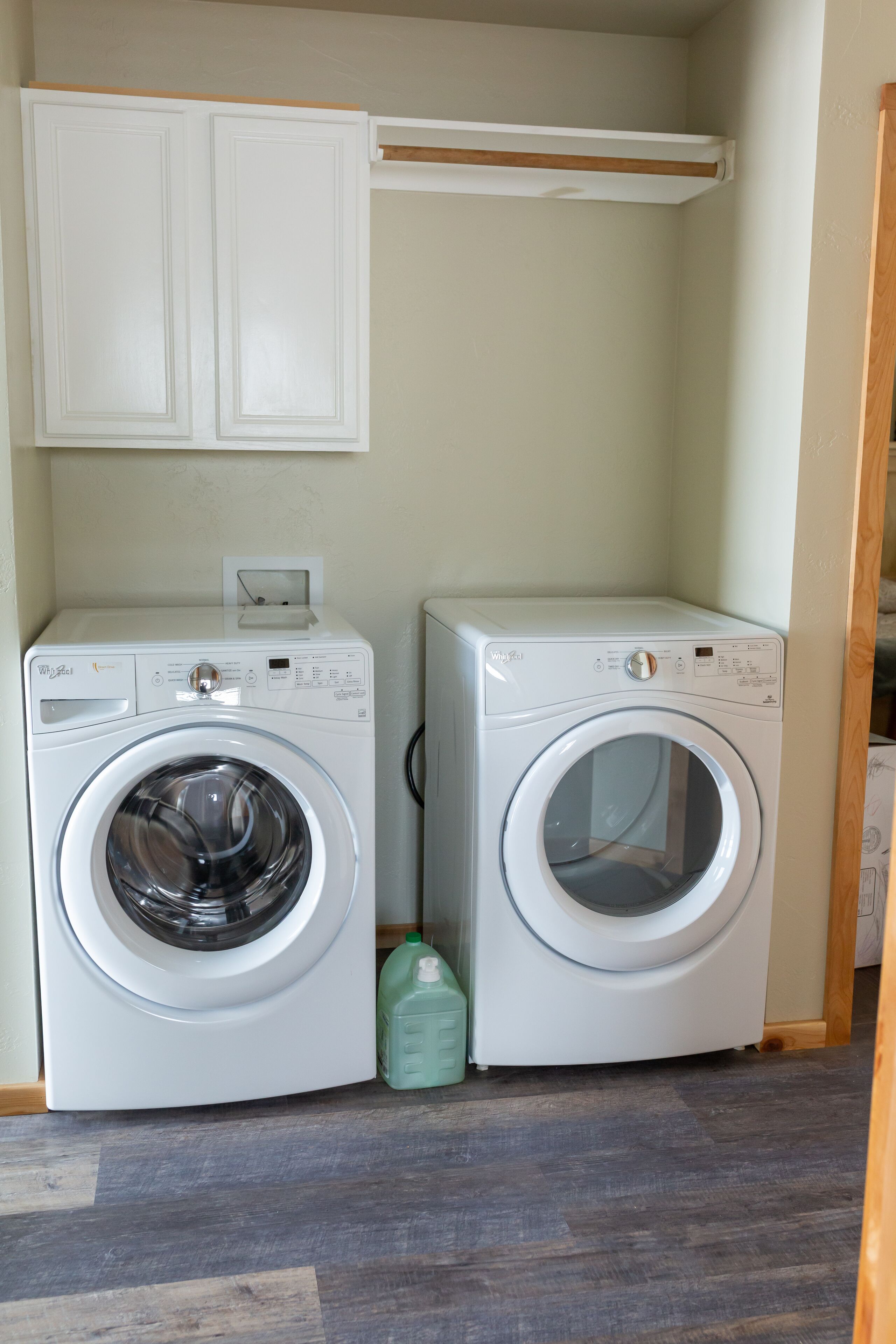 A laundry room with two white front-loading washing machines, a green detergent bottle, and a wooden shelf above the machines.