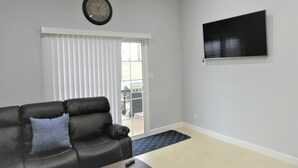 A living room with a black leather sofa, a blue pillow, a clock, a sliding glass door, and a flat-screen TV mounted on the wall. - Spacious Home* 7 minutes to Disneyland (Anaheim)