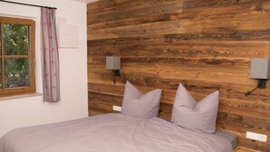 Ein Schlafzimmer mit einem Holz-Kopfteil, einem Bett mit einem hellvioletten Überwurf, zwei Wandlautsprechern, einem Fenster mit Vorhängen und einem Nachttisch mit Schublade.