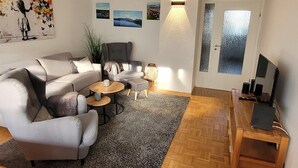 A modern living room with a grey sofa, a coffee table, and a TV stand. There are wall art pieces and a large window. - °Ferienwohnung Hedy mit 1 sep. Schlafzimmer, Südterrasse, 55 qm (Prien am Chiemsee)