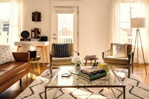 A living room with a brown leather sofa, a glass coffee table with books, a patterned rug, and a view through a door.