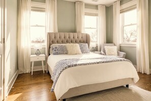 A bedroom with a large bed, a nightstand, a chair, and a window with curtains.