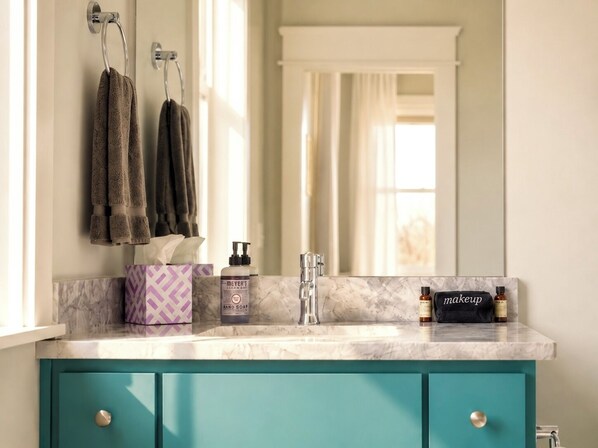 Un baño con encimera de mármol, un mueble de color aguamarina, un espejo y artículos de aseo personal, incluyendo un dispensador de jabón y una bolsa de maquillaje.