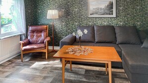 Una sala de estar con un sillón de cuero, un sofá, una mesa de centro de madera con un centro de mesa floral, una lámpara de pie y un cuadro de un paisaje en la pared.