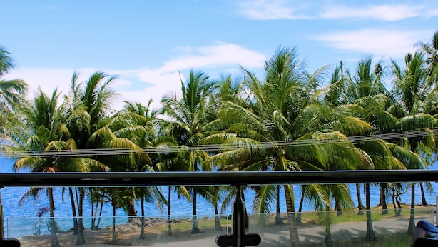 A balcony with a view of palm trees and the ocean. There is a table and two chairs.
