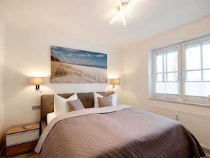 Une chambre avec un grand lit, deux lampes de chevet, une fenêtre et un tableau représentant une scène de plage accroché au mur.