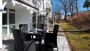 En udendørs terrasse med et bord og stole, en hvid bygning med altaner, og et græsstykke med træer.