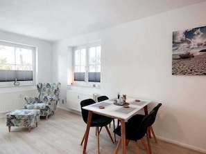 Un coin repas avec une table blanche et des chaises noires, un fauteuil à motifs et un tableau représentant une scène de plage accroché au mur.