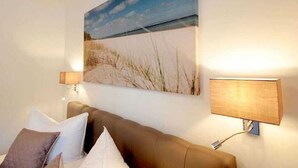 Et soveværelse med et stort maleri på væggen af et strandmotiv, to sengeborde med lamper, og en brun sofa med puder.
