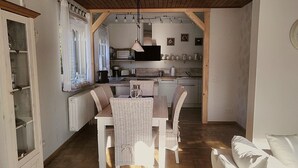 Een gezellig eetgedeelte met een houten tafel en rieten stoelen, een keuken op de achtergrond en een raam met gordijnen.