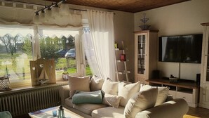 Een gezellige woonkamer met een beige bank, een bijzettafeltje met een boek, een televisie en een raam met gordijnen.