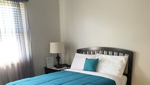 A neatly made bed with a teal bedspread and white pillows, a wooden headboard, a bedside table with a lamp, and a window with sheer curtains.