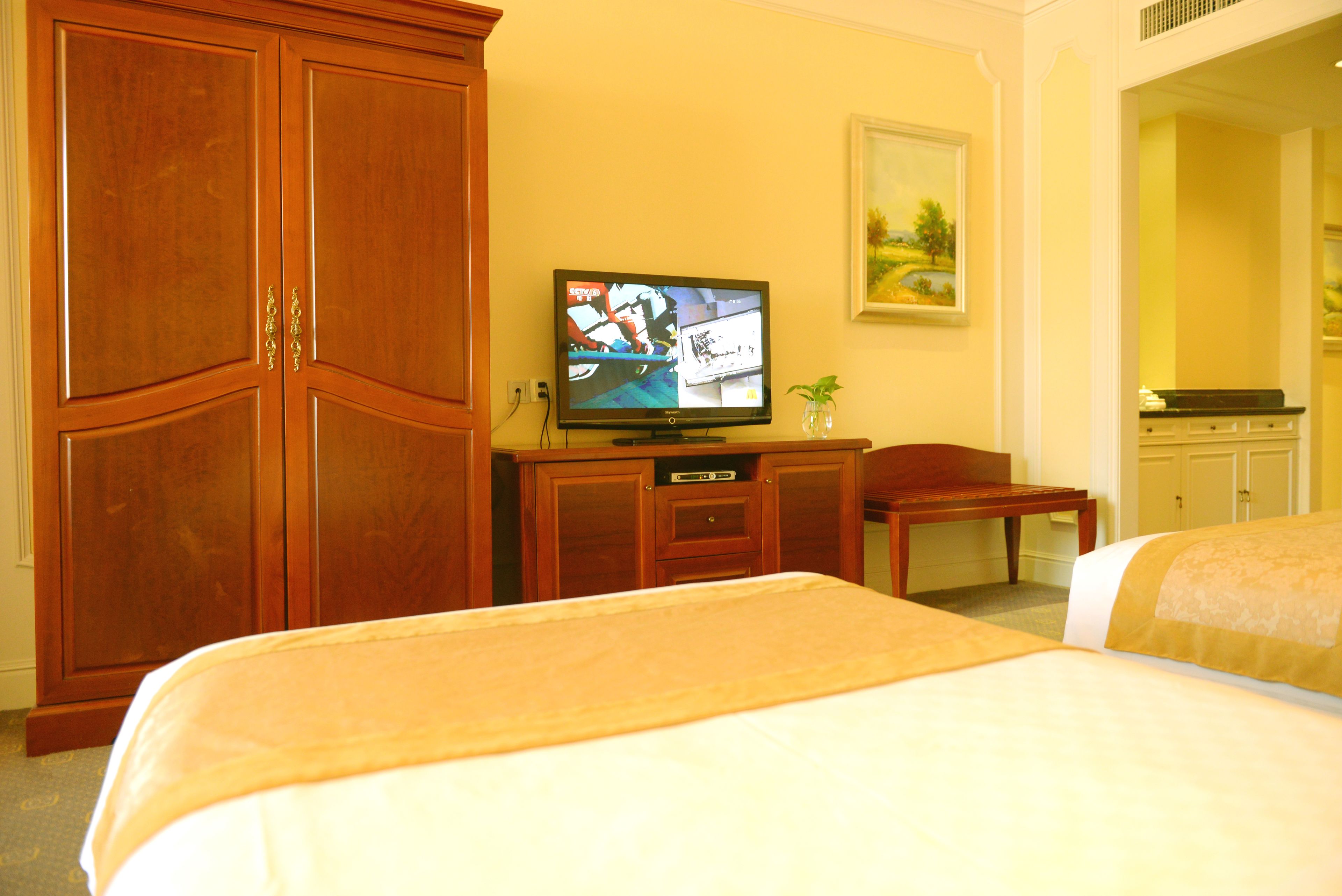 A hotel room with a large wooden wardrobe, a television on a wooden cabinet, a small table with a chair, and two beds with beige bedding.