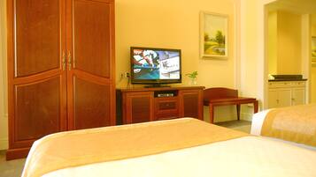 A hotel room with a large wooden wardrobe, a television on a wooden cabinet, a small table with a chair, and two beds with beige bedding.