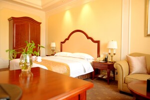 Habitación de hotel con una cama grande, mesitas de noche, un armario de madera, una zona de estar con una silla y una mesita con un jarrón.