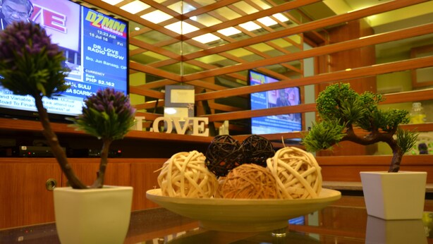 A reception area with a wooden reception desk, a television displaying a program, and a decorative plate with spherical objects on a glass table. - A Homey Place Manila, 1 Bedroom (Manila)