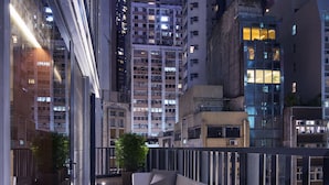 A modern outdoor seating area with a sofa and a table, a vase with flowers, and a cityscape view at dusk.