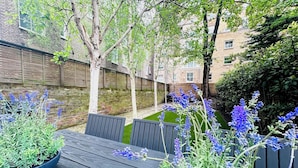 An outdoor patio with a wooden table and chairs, surrounded by blue flowering plants and trees. - Notting Hill Newly Decorated 3-Bedrooms with large Private Garden (London)