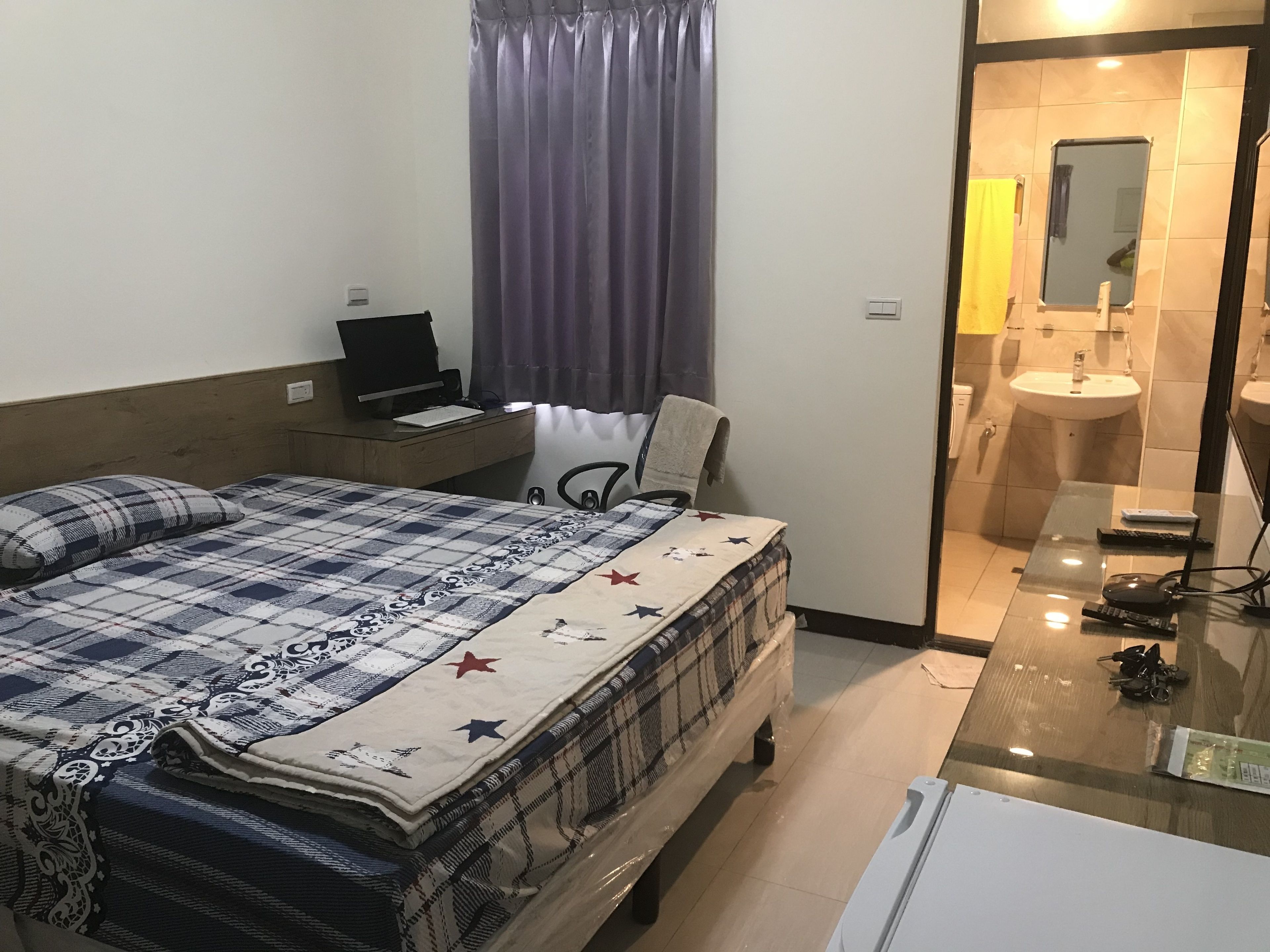 A hotel room with a bed, a desk with a laptop, a chair, a bathroom with a sink and mirror, and a glass table with keys and a remote control.