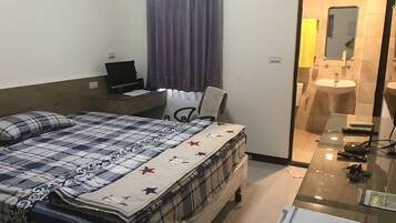 A hotel room with a bed, a desk with a laptop, a chair, a bathroom with a sink and mirror, and a glass table with keys and a remote control.
