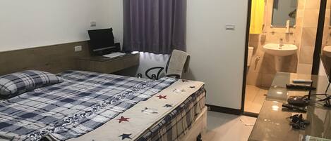 A hotel room with a bed, a desk with a laptop, a chair, a bathroom with a sink and mirror, and a glass table with keys and a remote control.