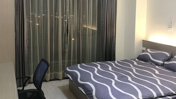 Een hotelkamer met een bed, een bureau, een airconditioner en uitzicht op de stad door dunne gordijnen.