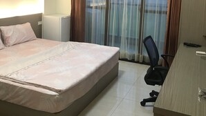 Een hotelkamer met een bed, bureau, airconditioning en uitzicht buiten door dunne gordijnen.