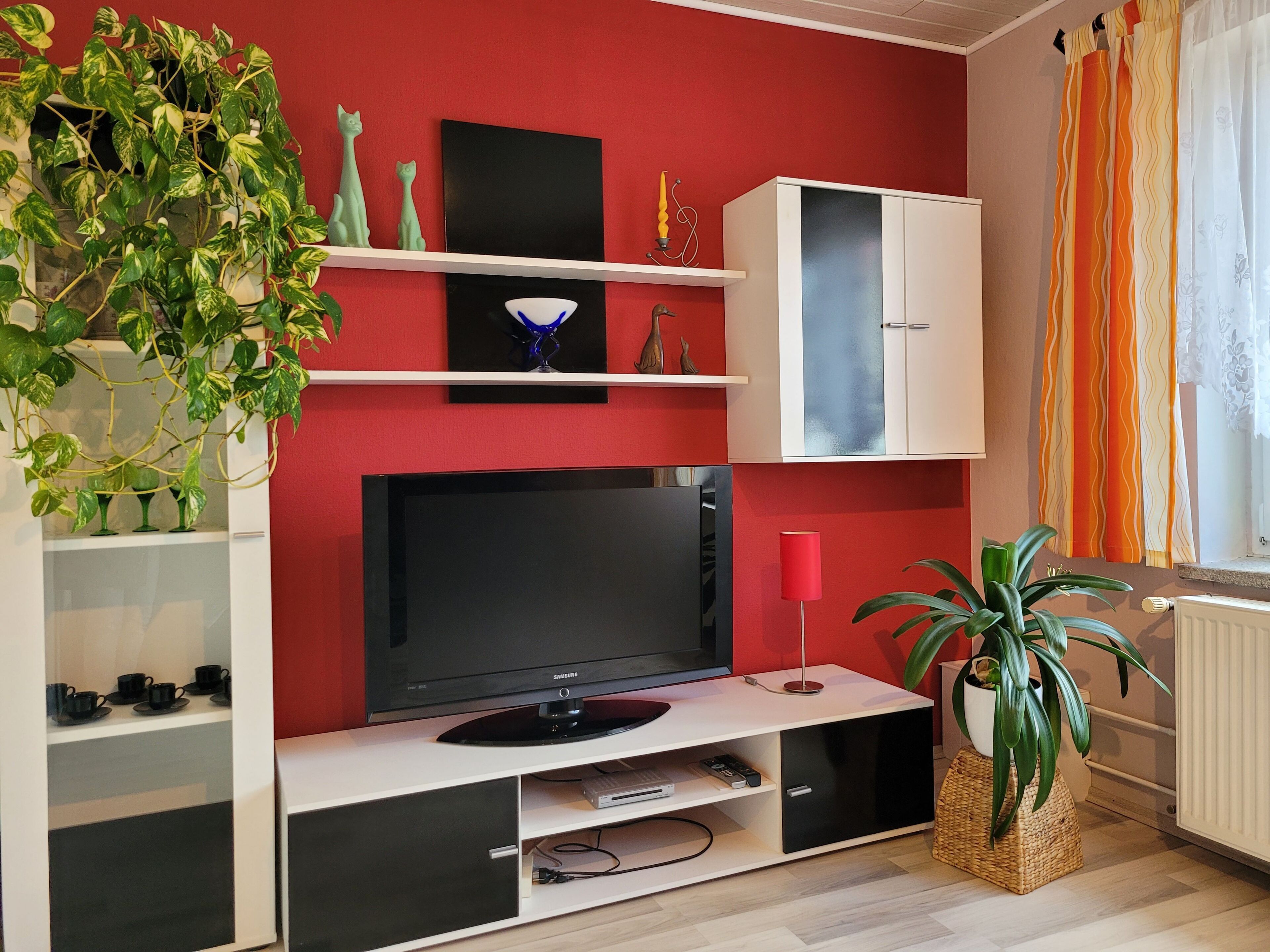 A modern living room with a red accent wall, a flat-screen TV, white shelving, and decorative items.