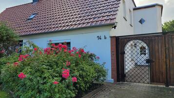 A house with a tiled roof, a number '14 b' on the wall, a wooden gate, and pink flowers in the garden.
