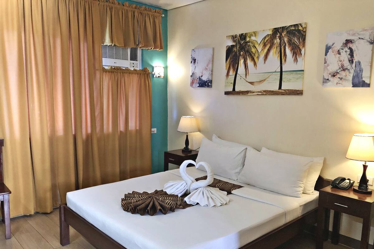 Une chambre d’hôtel comprenant un lit, des rideaux, un climatiseur fixé au mur, une lampe de chevet et des œuvres d’art représentant des scènes tropicales.