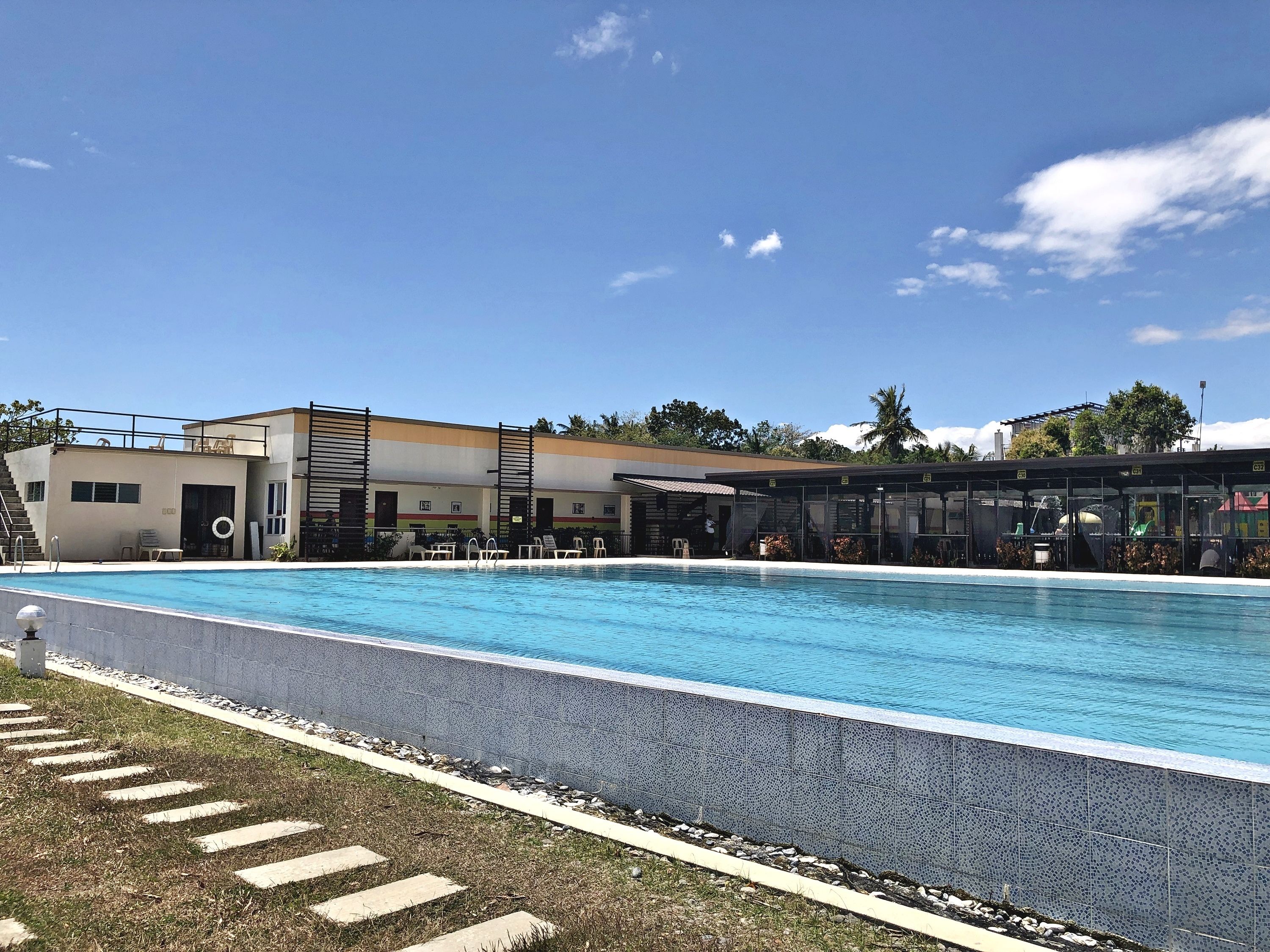Una piscina amb una zona de solàrium amb rajoles, una zona de seients coberta i un edifici amb teulada plana i grans finestres.