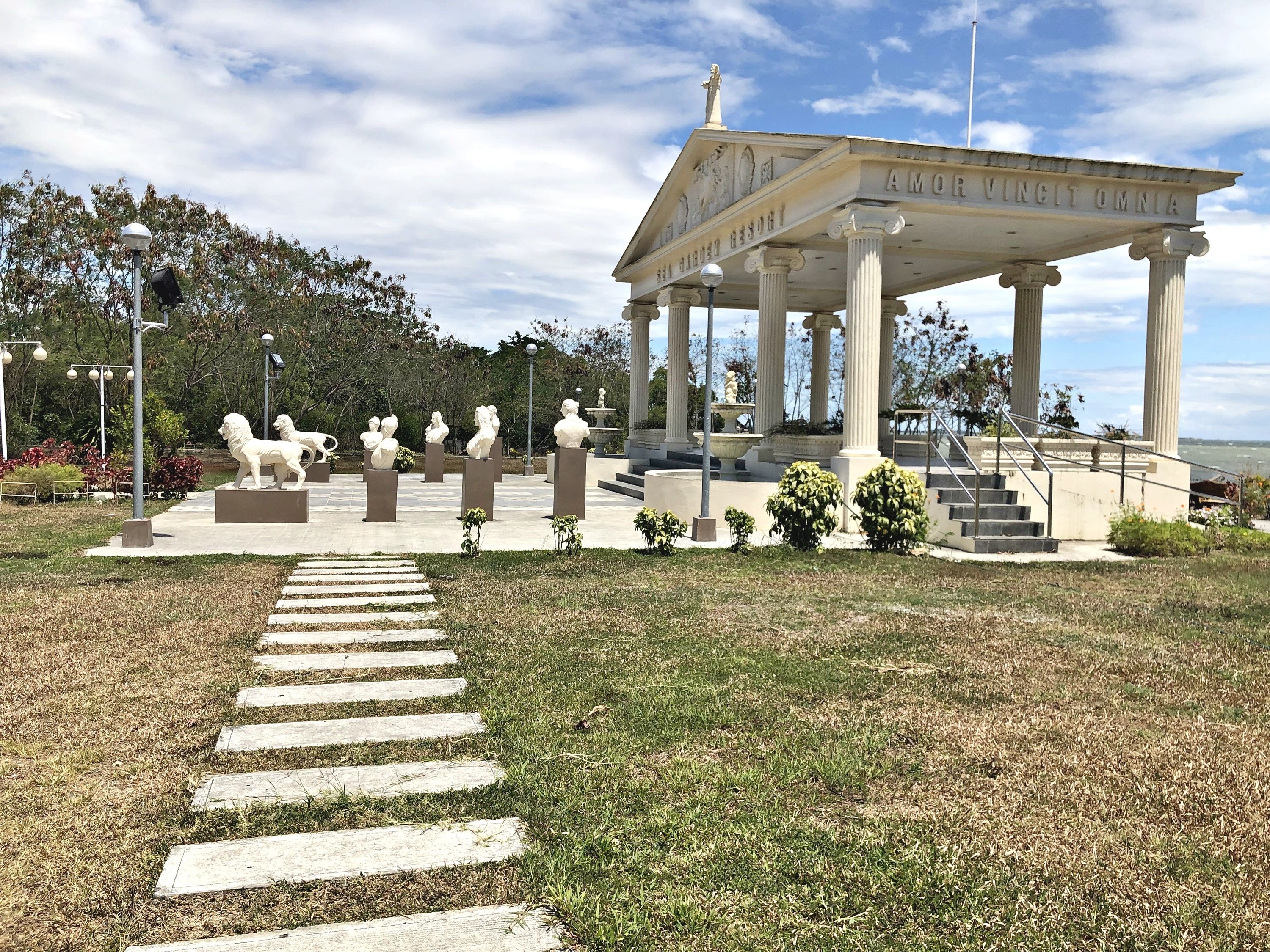 A pavilion with columns and a statue on top, surrounded by sculptures and a pathway.