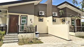 Uma casa moderna de dois andares com telhado plano, aparelhos de ar condicionado, porta de entrada e uma pequena varanda com cadeiras de plástico.