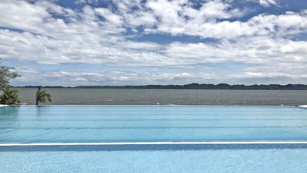 Piscina azul e límpida com vista para o mar e colinas ao longe, sob um céu parcialmente nublado.