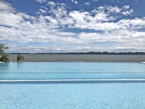 Une piscine d’un bleu éclatant, offrant une vue sur la mer et les collines lointaines, sous un ciel partiellement nuageux.