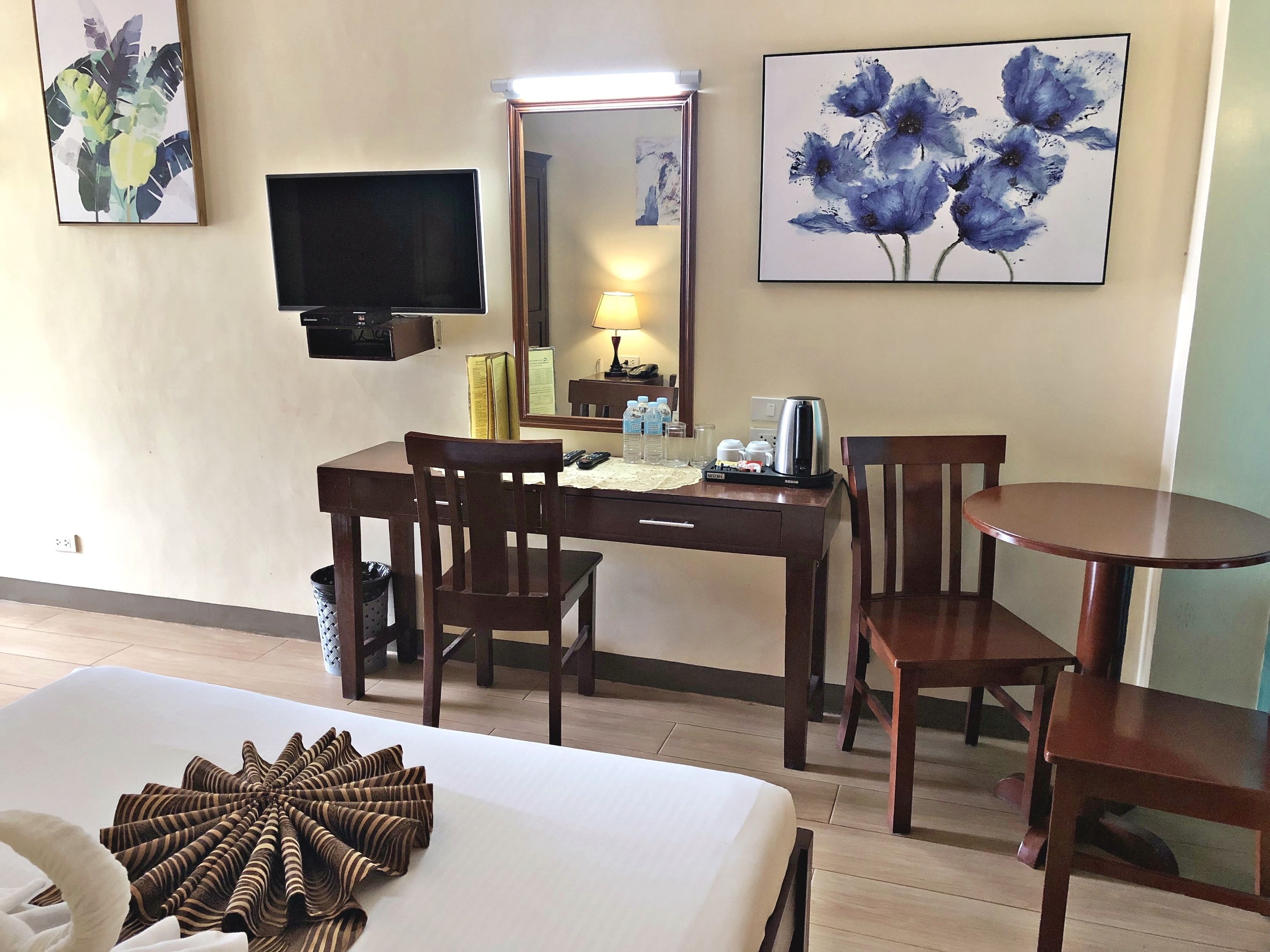 Habitación de hotel con cama, escritorio con silla, una mesa redonda y dos sillas. Hay un televisor en la pared, un espejo y obras de arte sobre el escritorio.