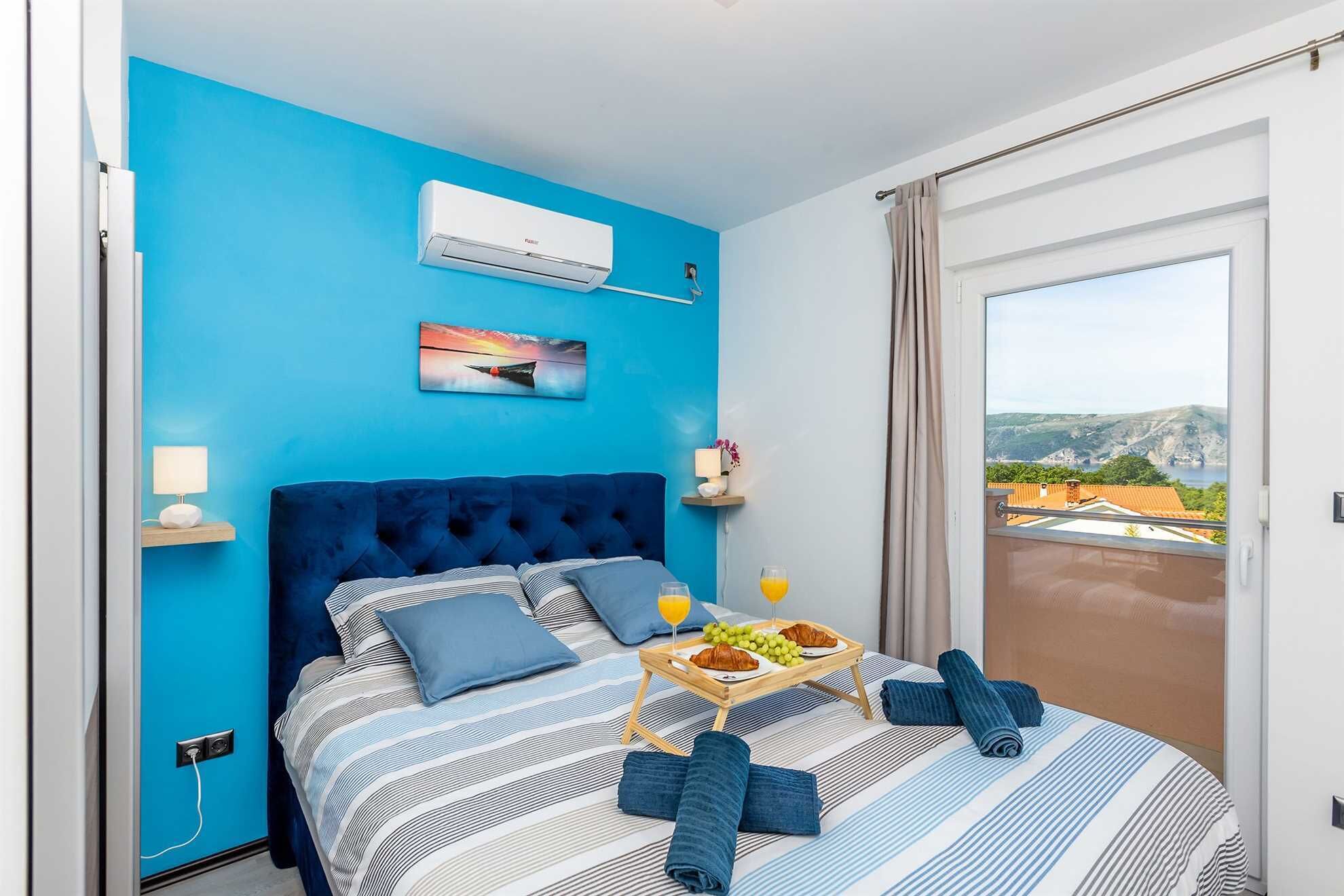 Une chambre d’hôtel avec un mur bleu, un grand lit avec une literie à rayures, une vue sur l’extérieur par une porte coulissante et un plateau de petit-déjeuner posé sur le lit.
