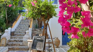 Un escalier en pierre mène à un bâtiment blanc doté d’une porte bleue, le tout étant entouré d’une végétation luxuriante et de fleurs roses éclatantes.