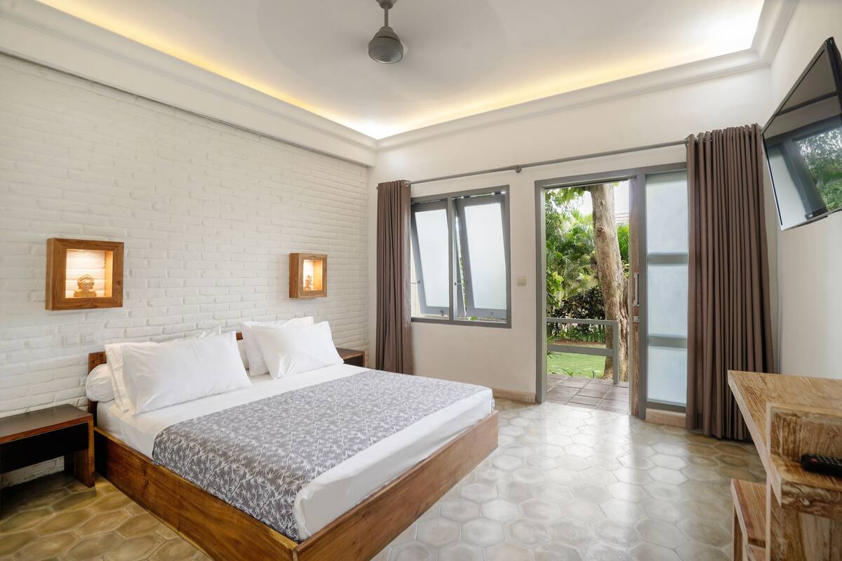 A bedroom with a large bed, a wooden nightstand, a desk with a chair, a TV mounted on the wall, and a sliding glass door leading to an outdoor area.