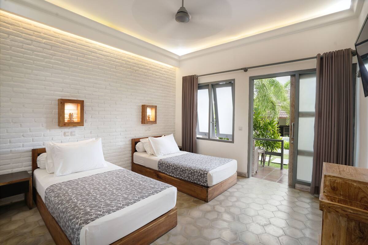 A hotel room with two beds, a white brick wall, and a large sliding glass door leading to an outdoor area.