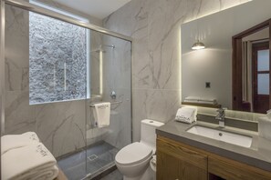 A modern bathroom with a glass shower enclosure, a marble-tiled wall, a sink with a mirror, and a toilet. - Hotel Casa Monarca (Puebla)