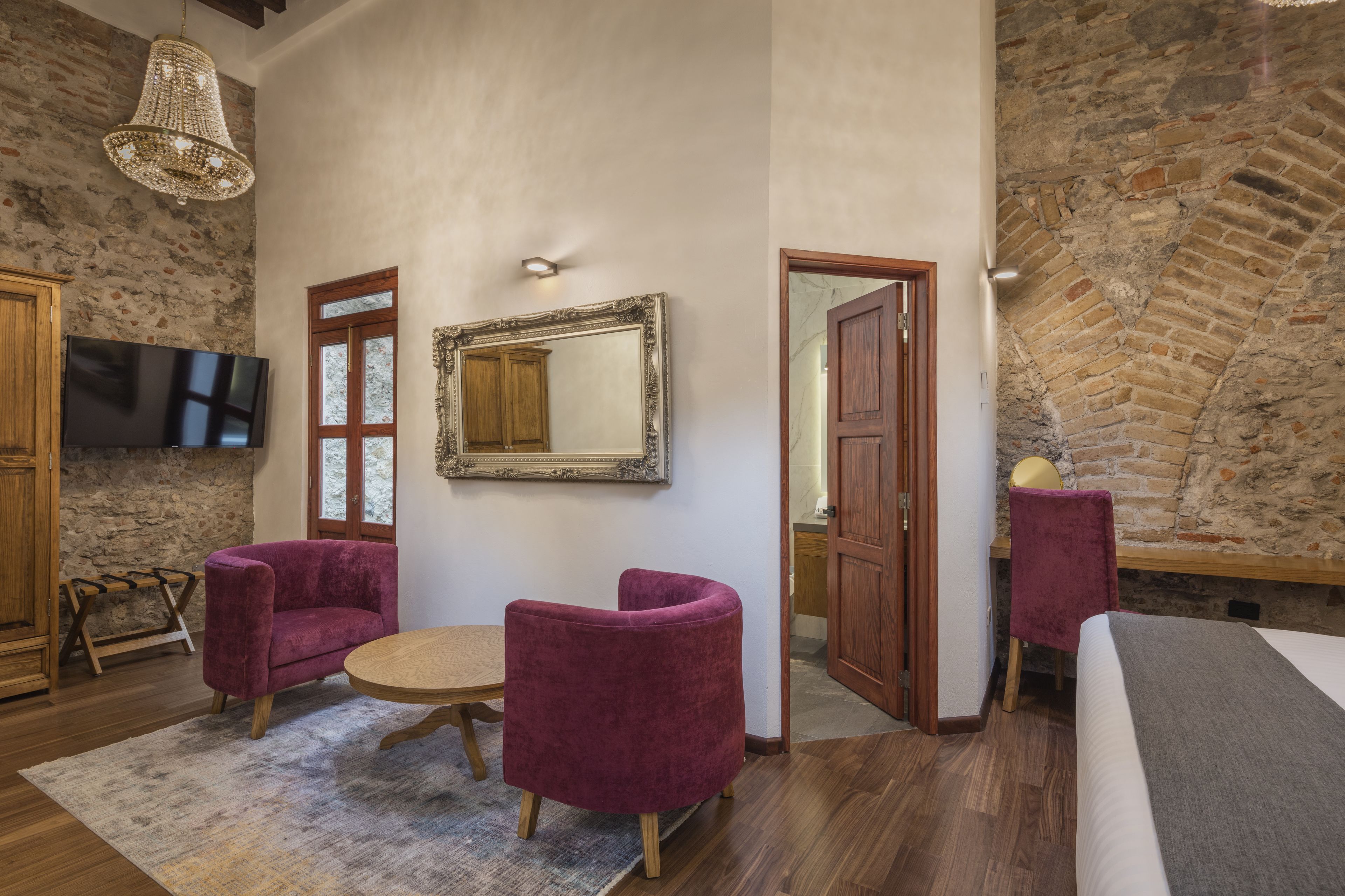 Une pièce avec un mur en pierre, une porte en bois, une table ronde, deux fauteuils violets, un téléviseur et un miroir.