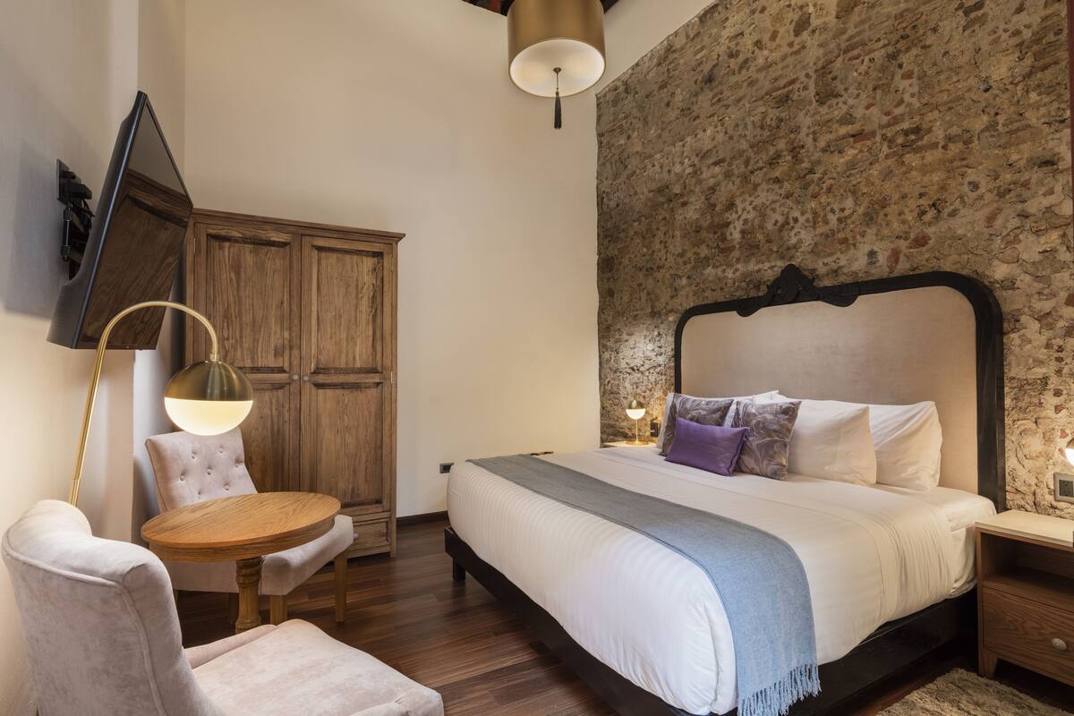 A bedroom with a bed, a round wooden table, a chair, a wardrobe, and a stone wall.