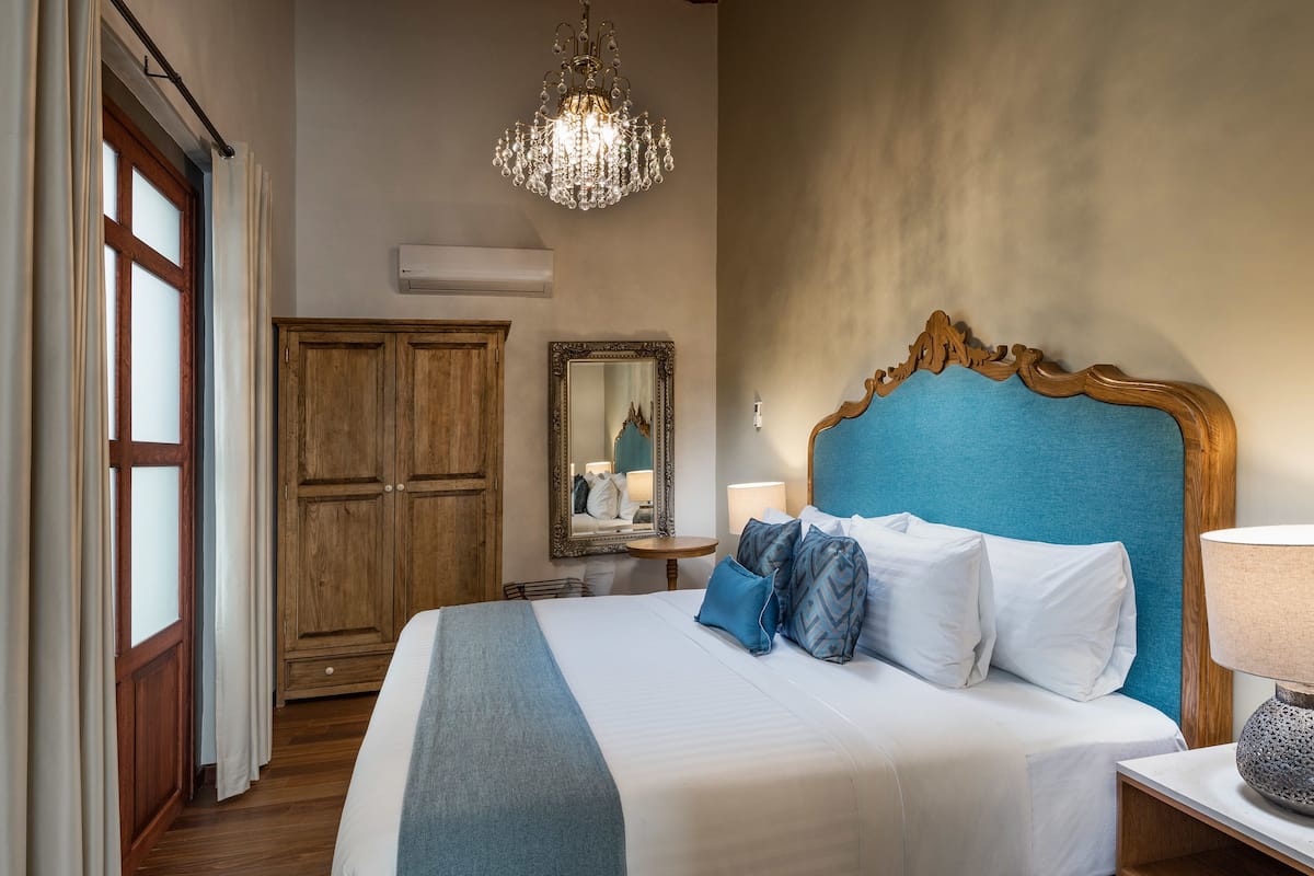 A neatly made bed with a blue headboard, a wooden wardrobe, a mirror, and a chandelier.