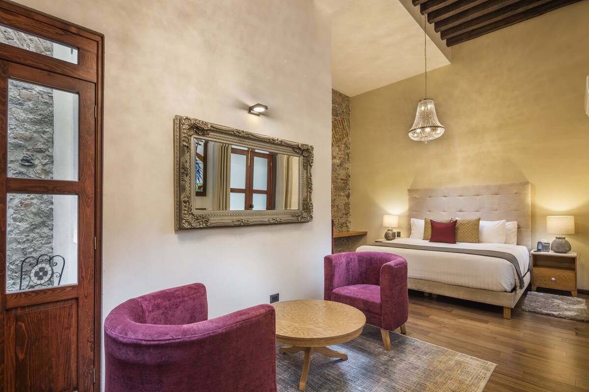 A bedroom with a bed, two purple chairs, a round table, a mirror, and a chandelier.